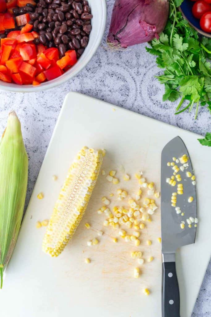 A cutting board with a partially cut ear of corn, a knife, corn kernels, an unshucked corn, and bowls of chopped vegetables and beans nearby.