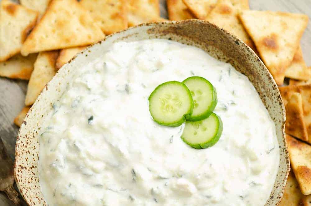 A bowl of tzatziki dip garnished with cucumber slices, surrounded by pita chips.