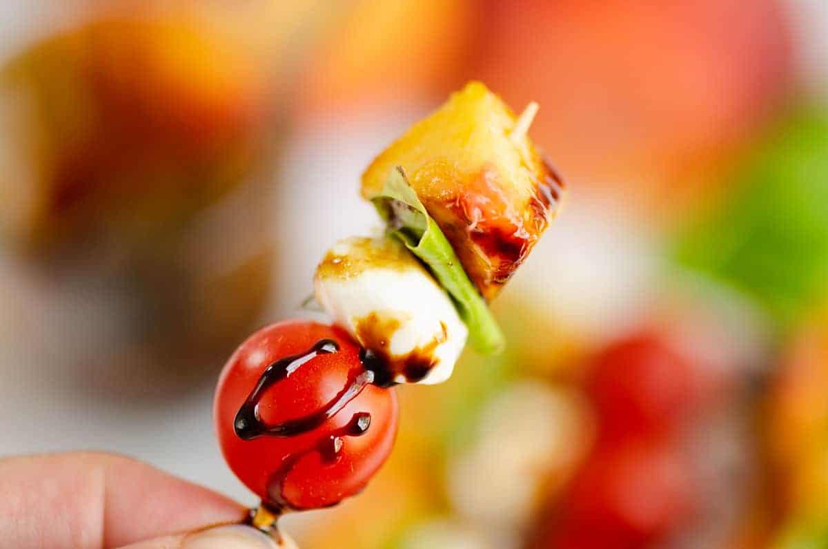A hand holds a skewer with a cherry tomato, mozzarella ball, basil leaf, grilled peach, and a drizzle of balsamic glaze.
