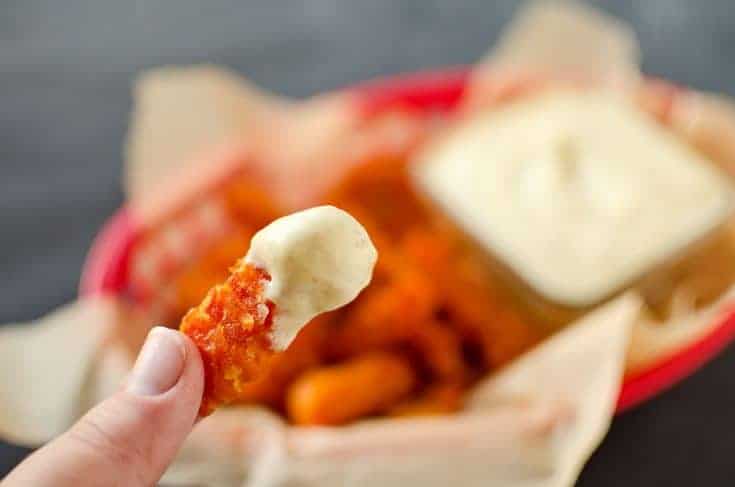 A hand holds a piece of breaded, fried food dipped in a creamy sauce, with more pieces and a sauce container in a red basket in the background.