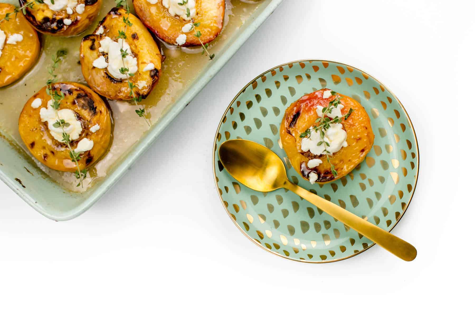 Baked peach halves topped with goat cheese and herbs on a green patterned plate with a gold spoon, next to a baking dish with more peaches.
