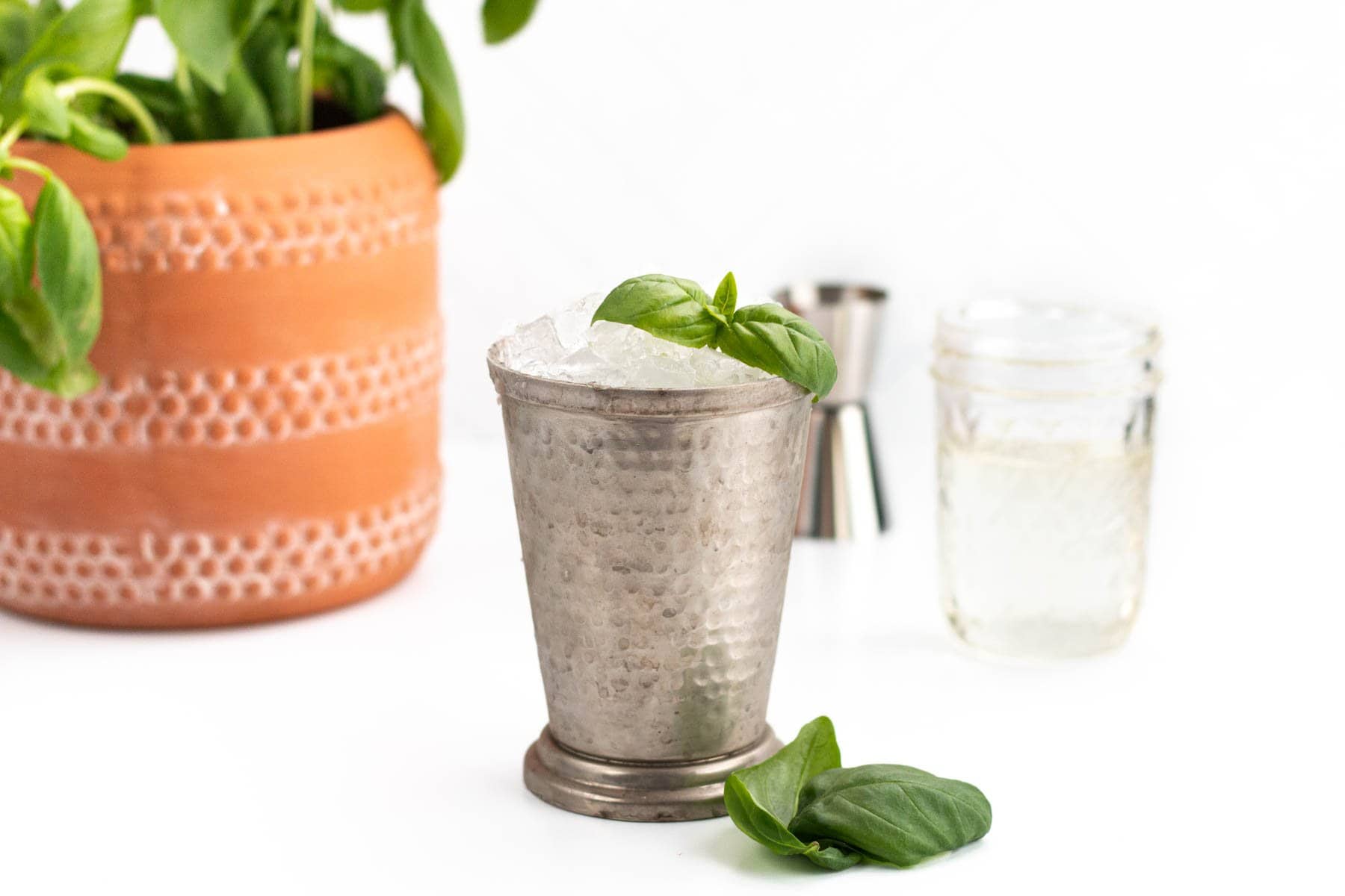 A metal cup filled with crushed ice and garnished with basil leaves, with a basil plant, jigger, and glass jar in the background.