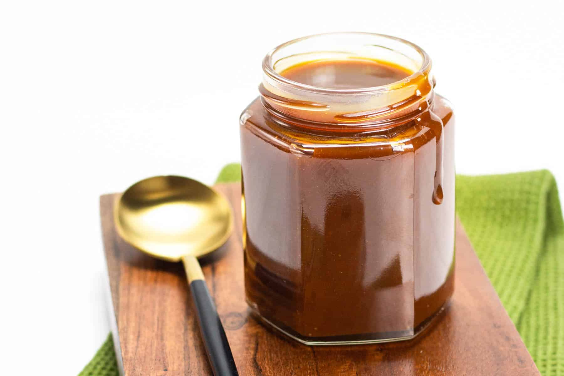 A jar of caramel sauce sits on a wooden board next to a gold and black spoon, with a green cloth in the background.
