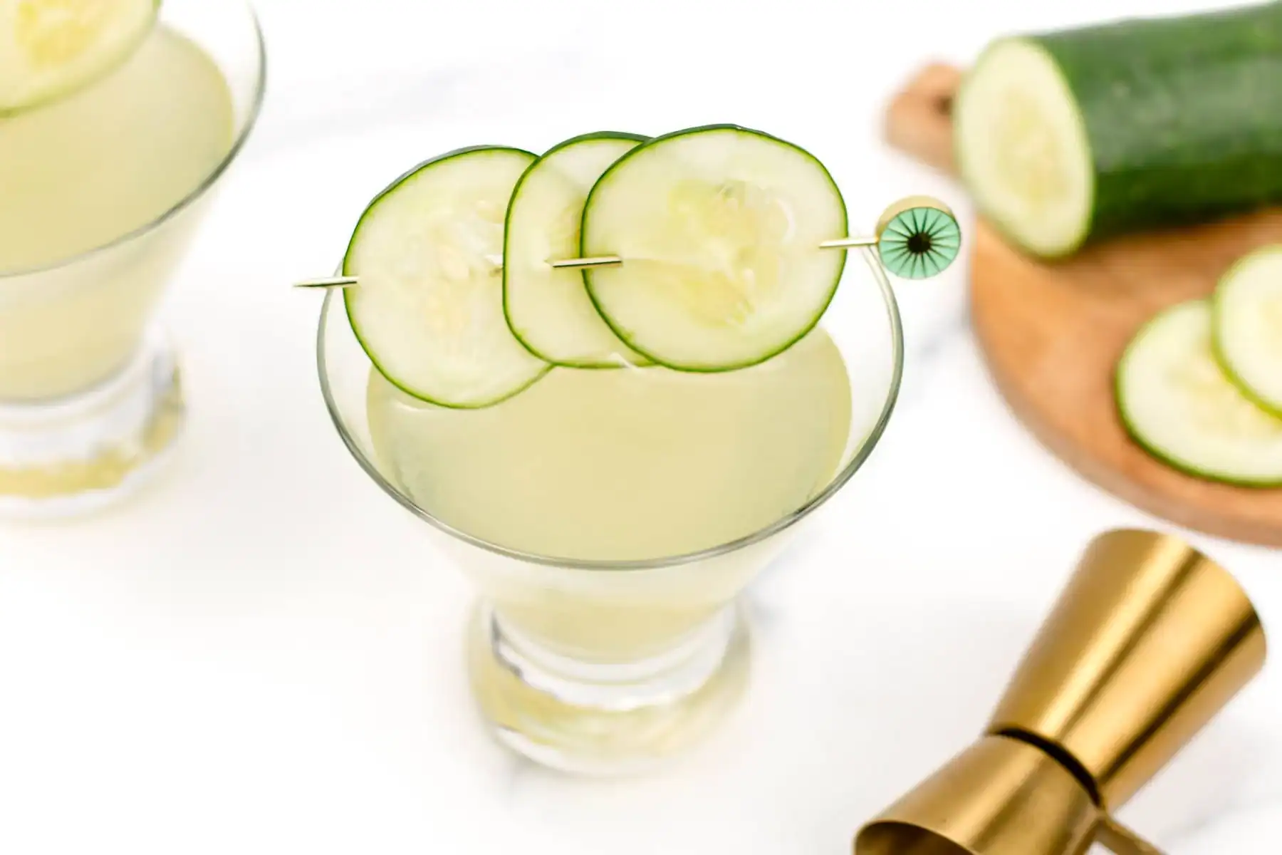 A cucumber cocktail garnished with cucumber slices on a skewer, with a jigger and sliced cucumber on a cutting board nearby.