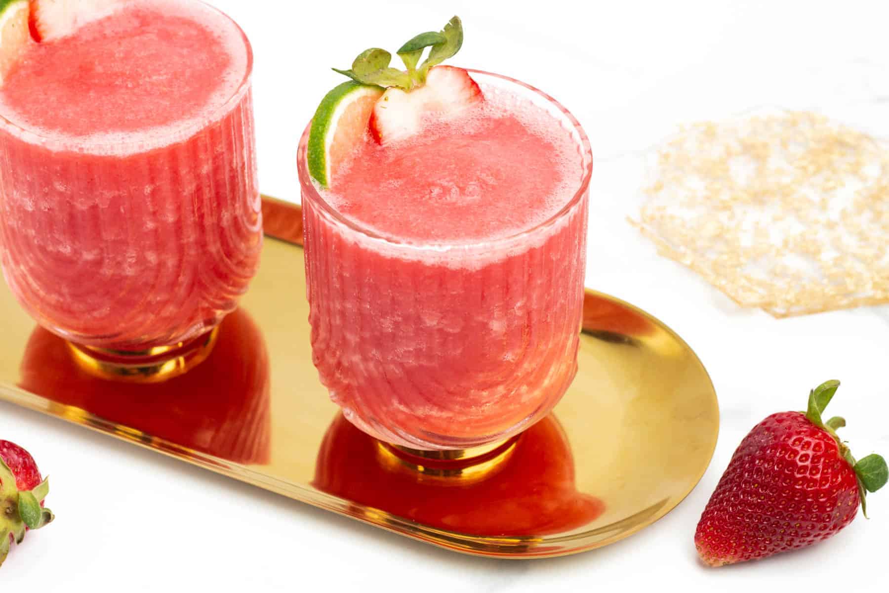 Two glasses of pink strawberry smoothie on a gold tray, garnished with strawberry slices, with whole strawberries and a pastry nearby.