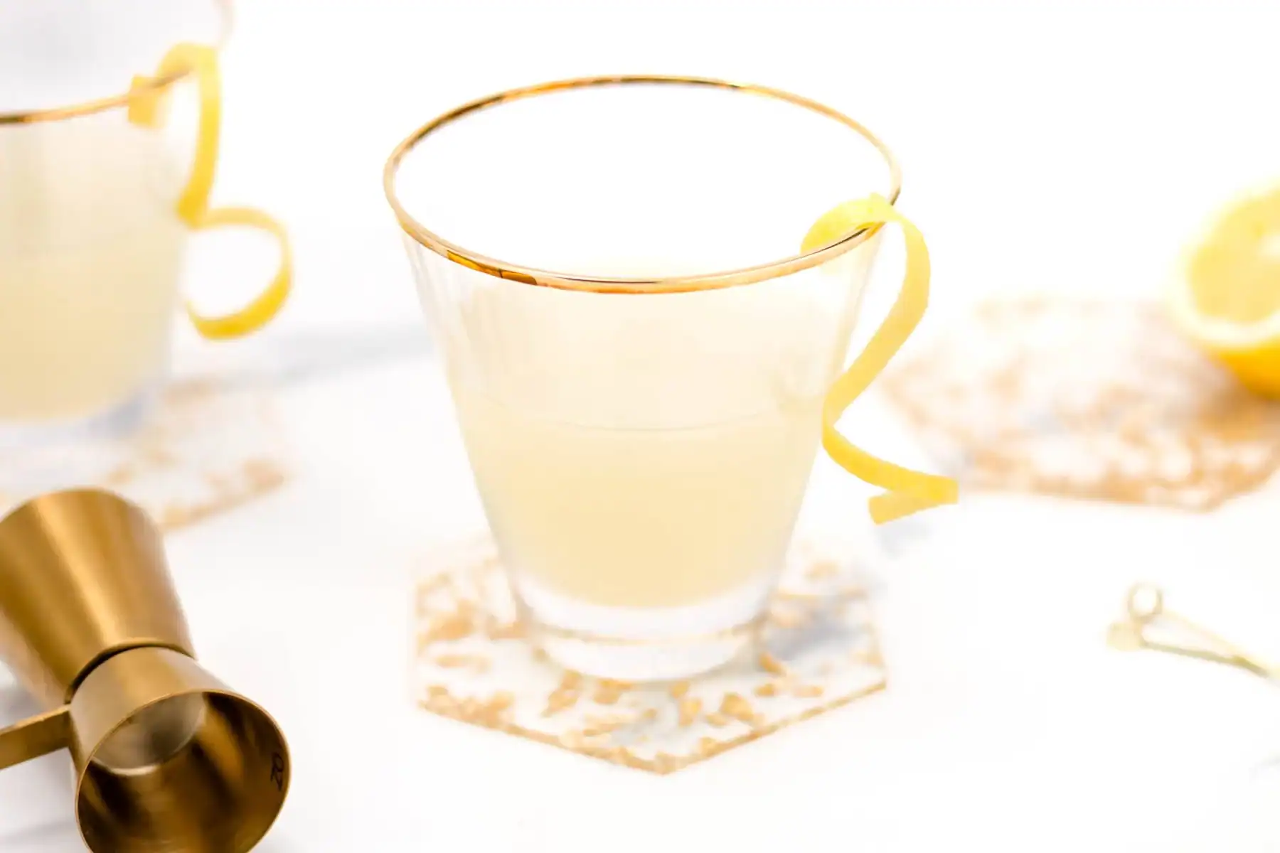 A clear glass with a gold rim containing a pale yellow drink, garnished with a lemon twist, sits on a hexagonal coaster next to a gold jigger and a lemon half.