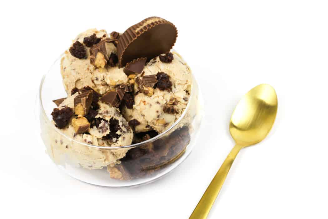 A glass bowl of ice cream topped with chocolate pieces and a peanut butter cup, next to a gold spoon on a white background.