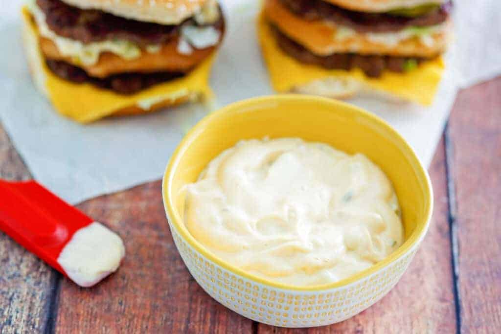 A yellow bowl filled with creamy white sauce sits on a wooden surface, with partially visible cheeseburgers and a red spoon nearby.