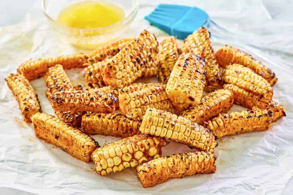 Grilled corn ribs are arranged on parchment paper with a small bowl of melted butter and a blue silicone brush in the background.