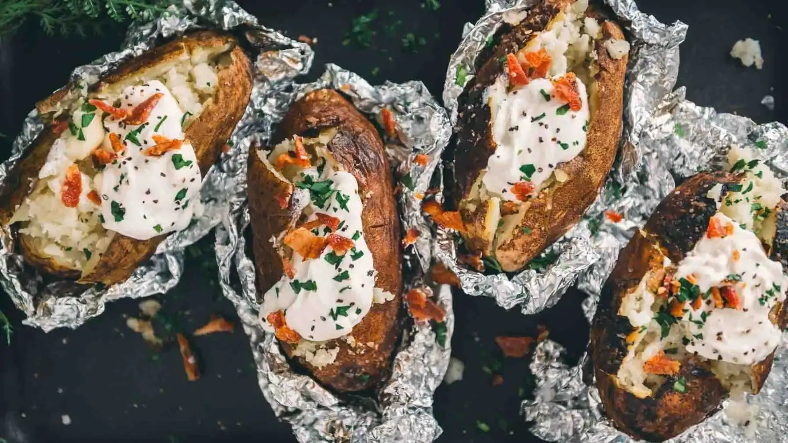 Four baked potatoes in foil topped with sour cream, chopped chives, and bacon pieces are arranged on a dark surface.