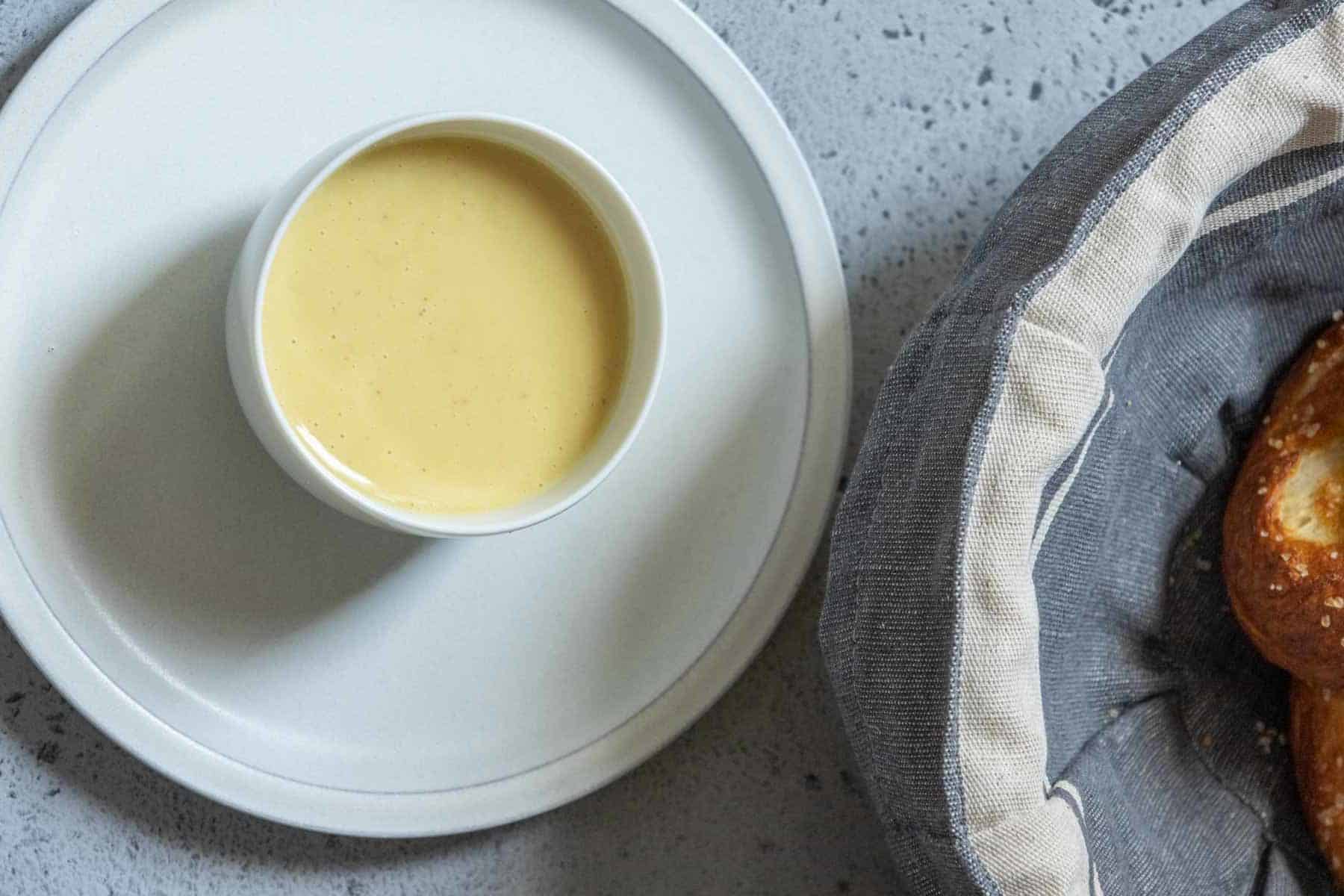 A small white bowl filled with creamy yellow sauce sits on a white plate next to a basket containing a pretzel.