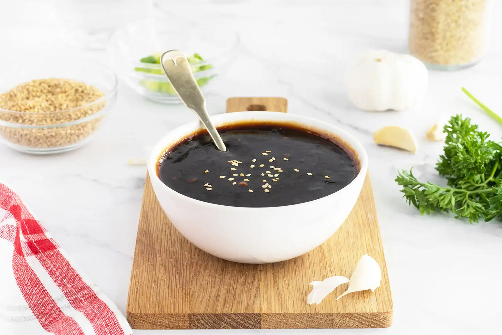 A white bowl filled with dark sauce, topped with sesame seeds, sits on a wooden board with a spoon; garlic, parsley, and other ingredients surround it.