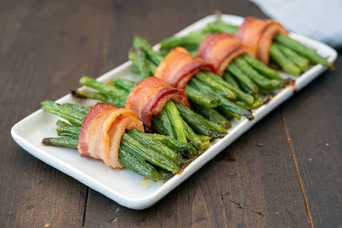 Five bundles of green beans wrapped in bacon are arranged in a row on a white rectangular plate, set on a dark wooden table.