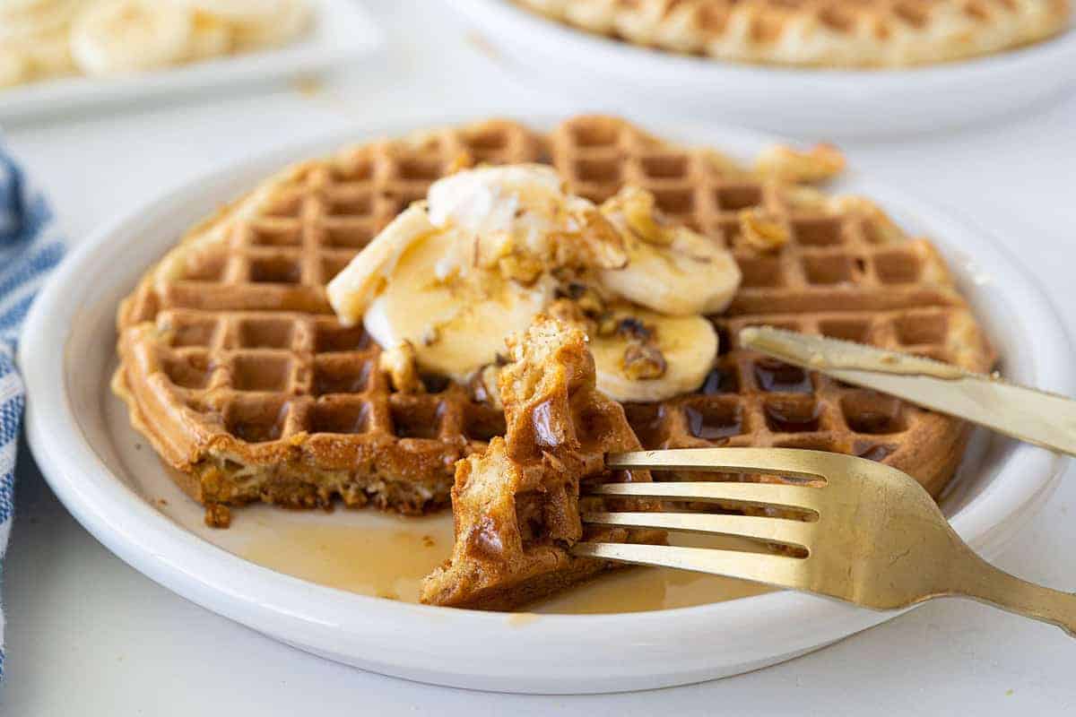 A plate with a waffle topped with banana slices and syrup. A fork and knife are cutting a piece from the waffle.