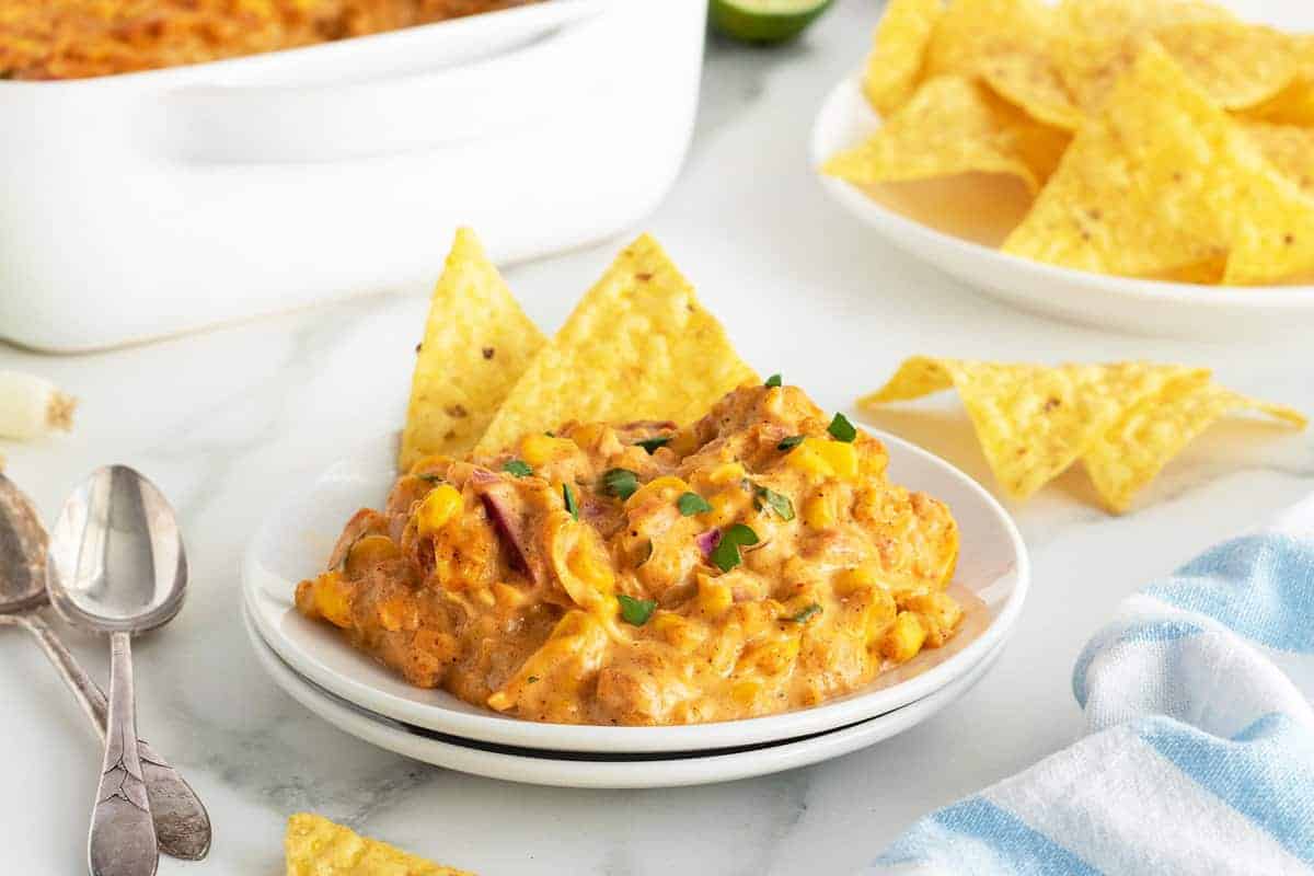 A plate of cheesy corn casserole with tortilla chips on a white plate, with more chips and a casserole dish in the background.