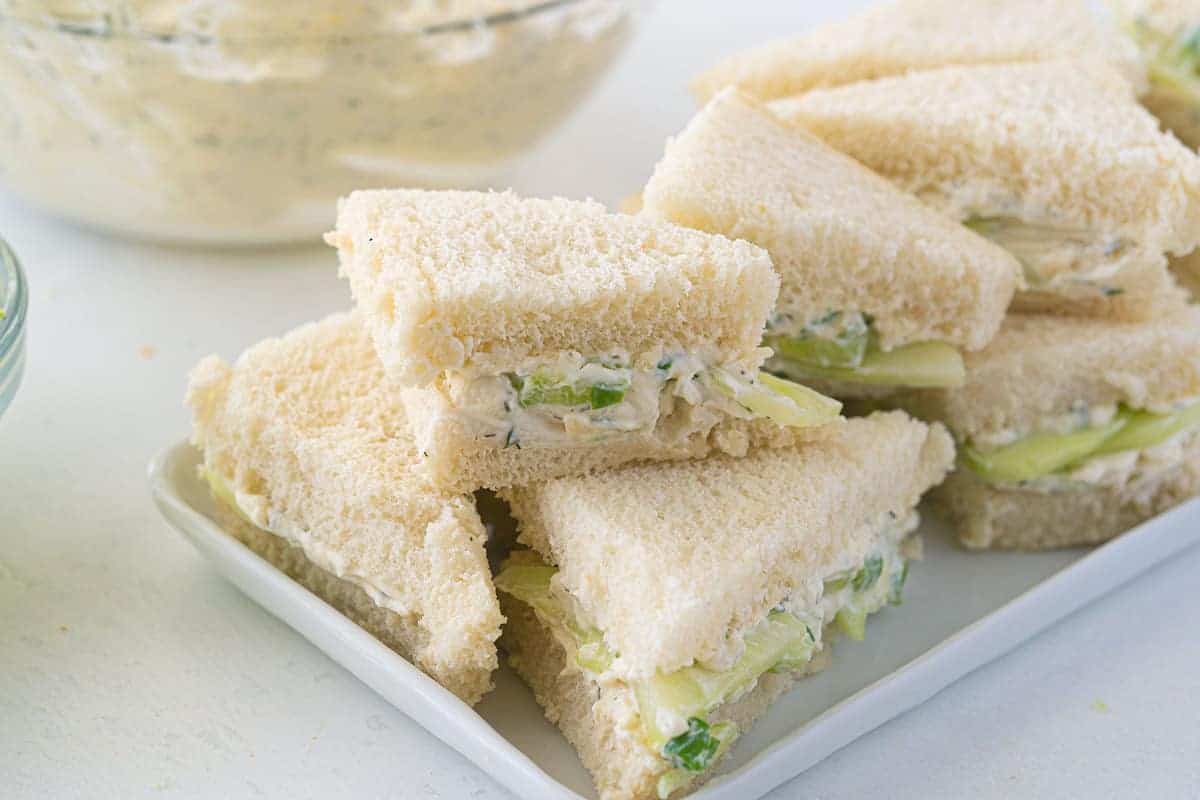 A plate of crustless white bread sandwiches filled with cucumber and a creamy spread, cut into triangles and stacked neatly.