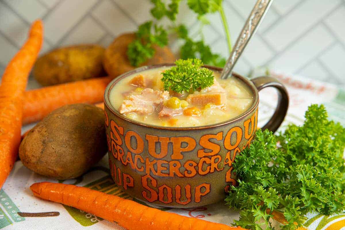 A mug of creamy soup with ham, corn, potatoes, and garnished with parsley, surrounded by carrots, potatoes, and fresh parsley on a kitchen counter.