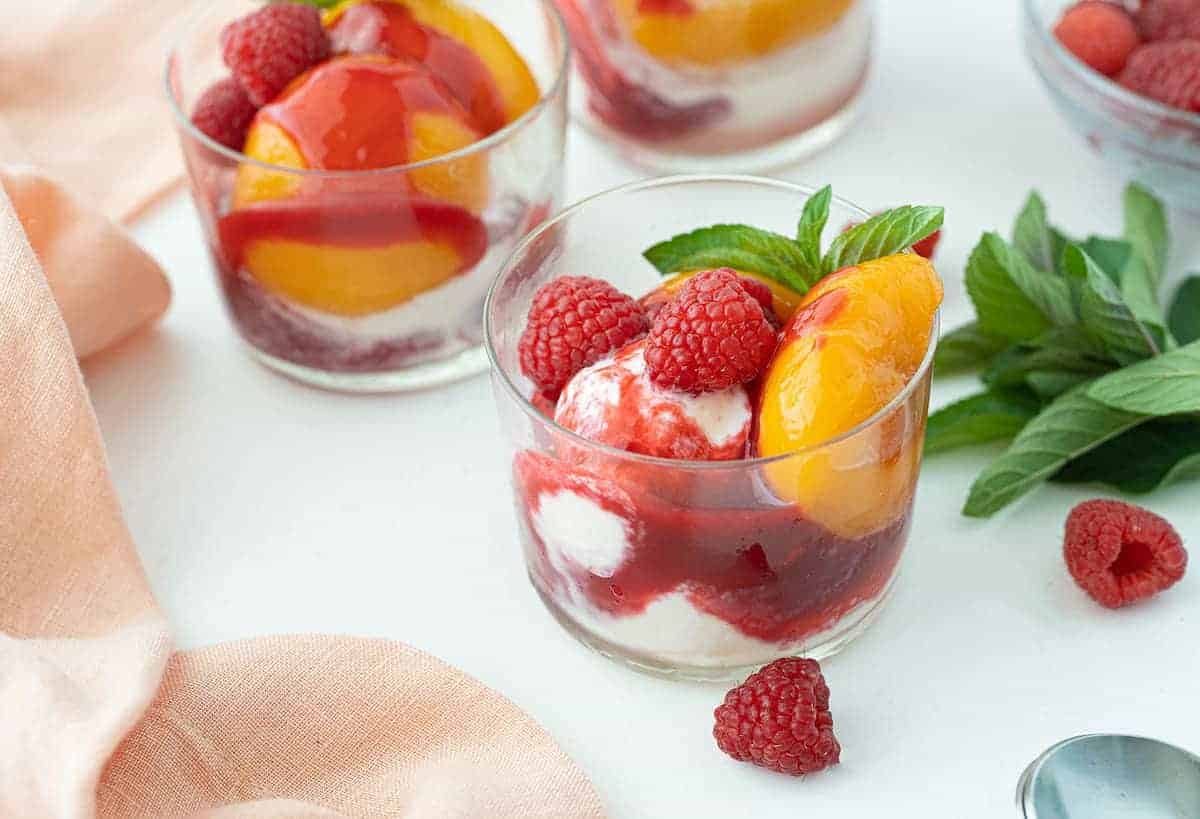 Three clear glasses filled with scoops of vanilla ice cream, peach halves, raspberry sauce, fresh raspberries, and mint leaves on a white surface.