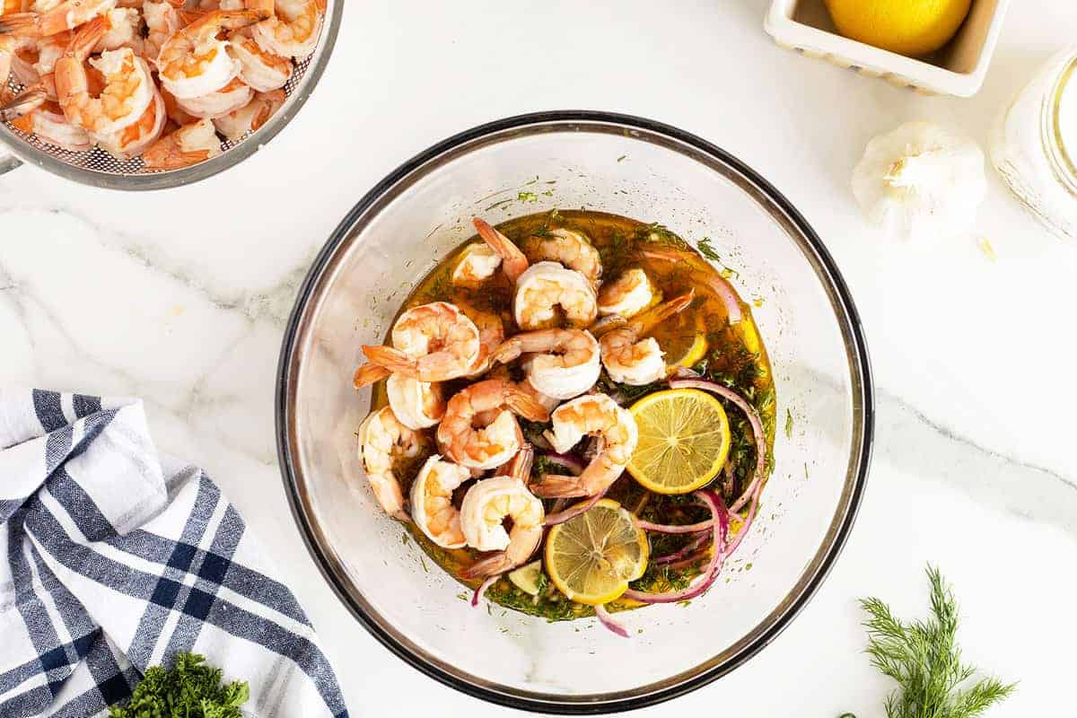 A glass bowl of shrimp marinating with lemon slices, red onion, and herbs on a white countertop, with additional shrimp and ingredients nearby.