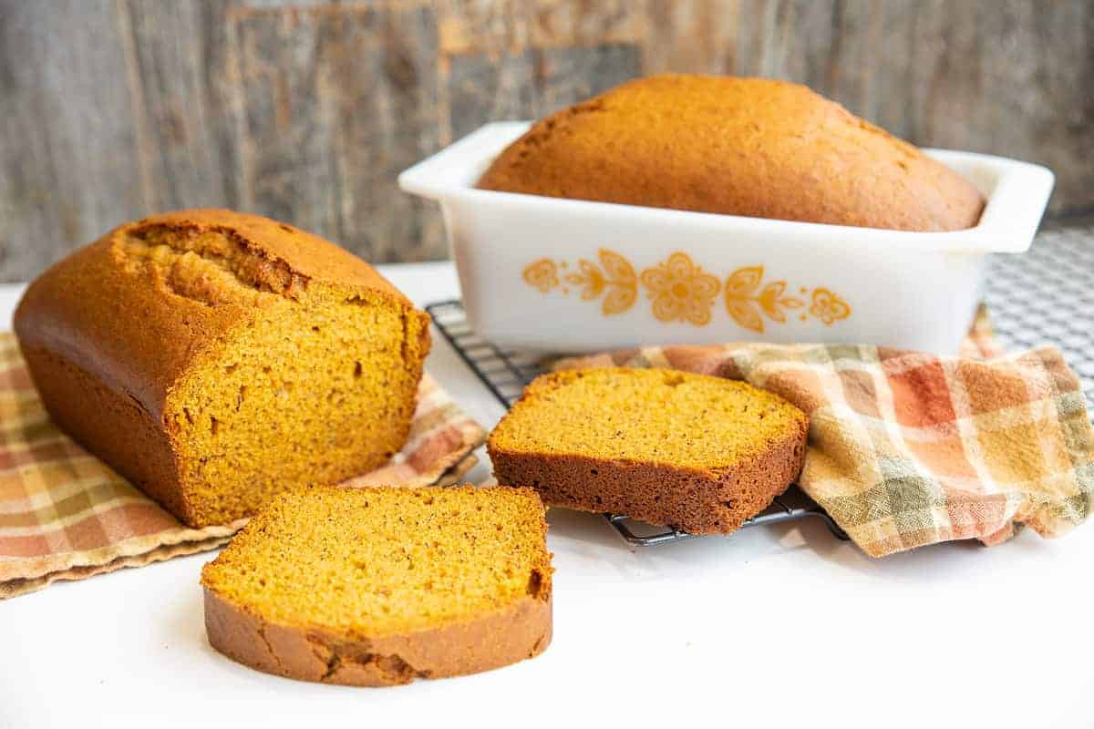 Two loaves of pumpkin bread, one sliced and one whole in a white baking dish, are displayed on a table with plaid cloths.