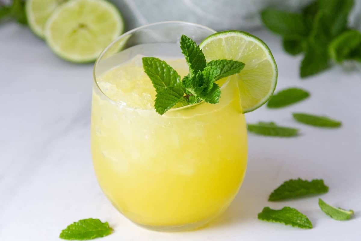 A glass of yellow slushy drink garnished with a lime slice and fresh mint leaves, with lime halves and mint scattered in the background.