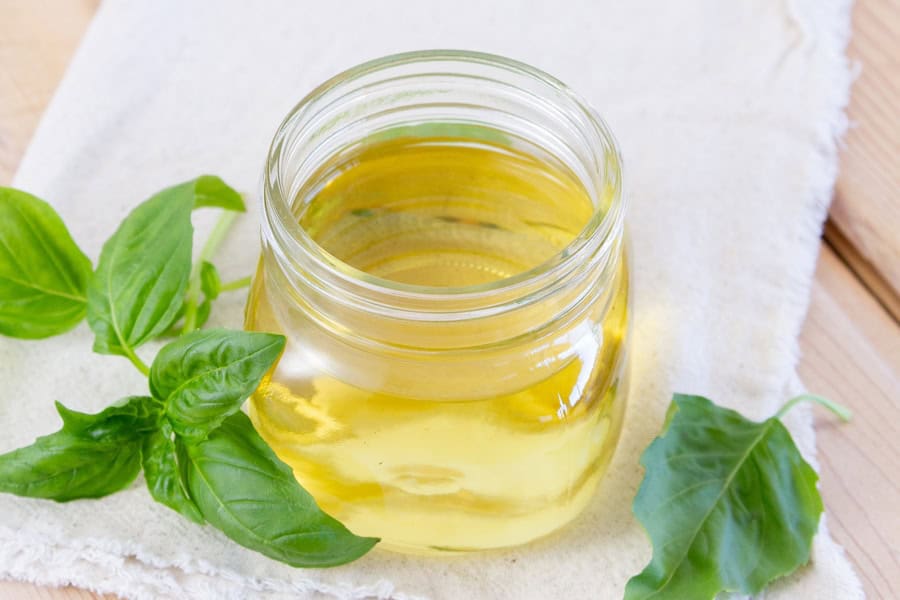 A glass jar filled with light yellow oil sits on a white cloth, with fresh green basil leaves placed beside it.