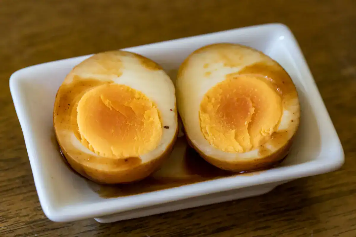 Two halves of a marinated soft-boiled egg are placed side by side in a small rectangular white dish on a wooden surface.