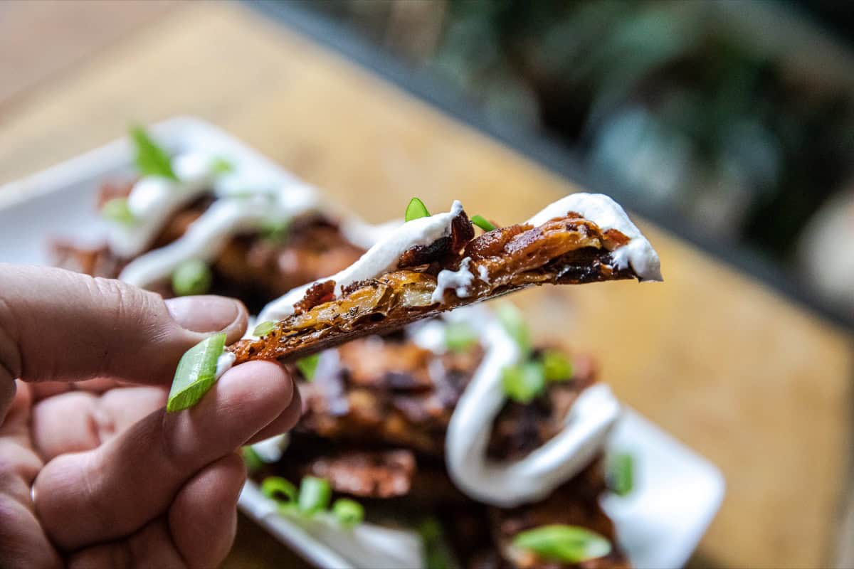 A hand holds a loaded potato skin topped with sour cream, bacon, and green onions, with more potato skins blurred in the background.