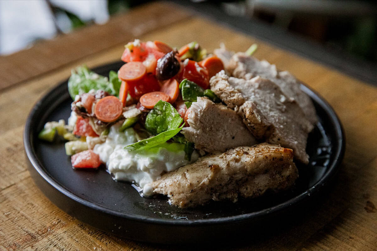 A black plate with sliced grilled chicken, a fresh salad with tomatoes, cucumbers, carrots, and cottage cheese, set on a wooden table.
