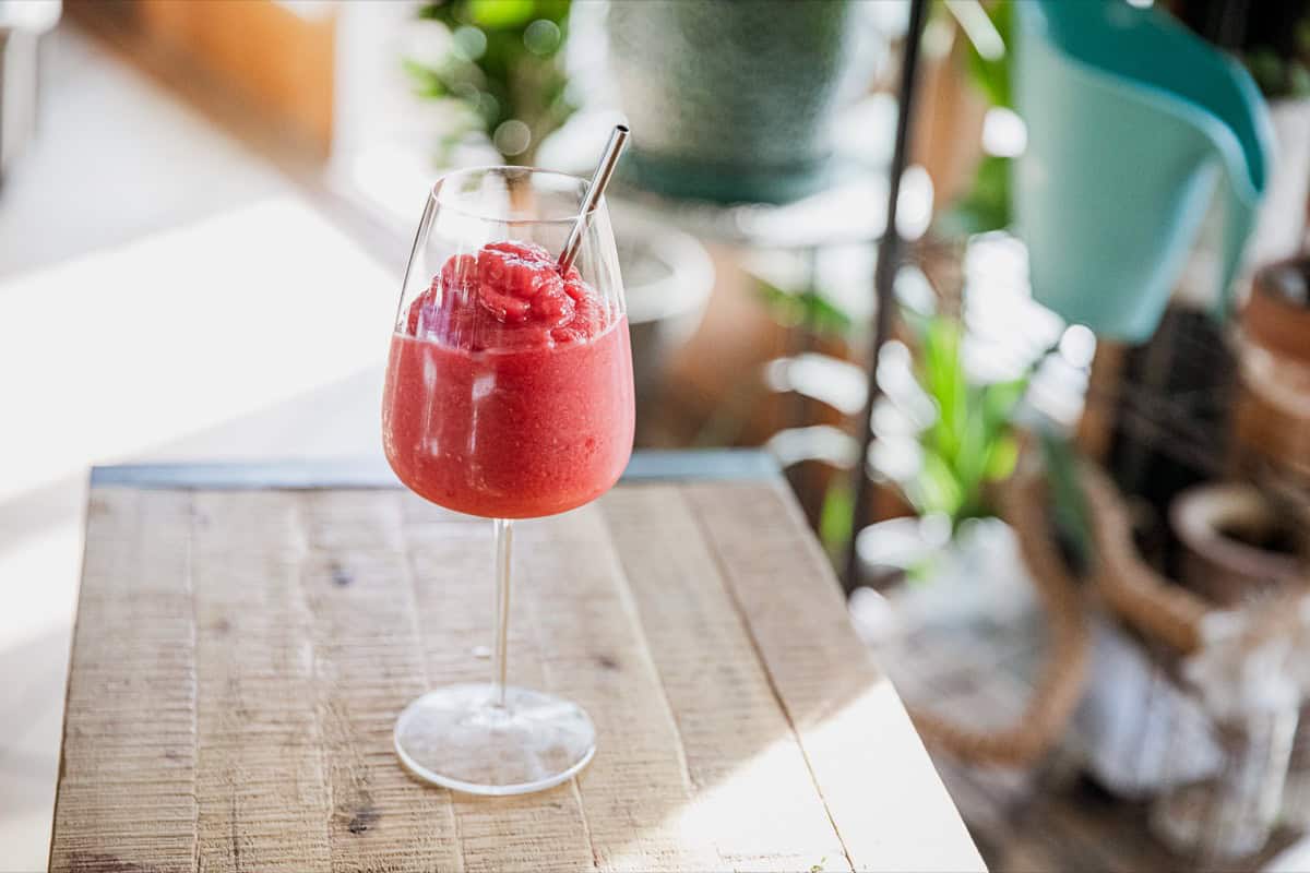 A glass of red frozen drink with a metal straw sits on a wooden table, with blurred plants and furniture in the background.