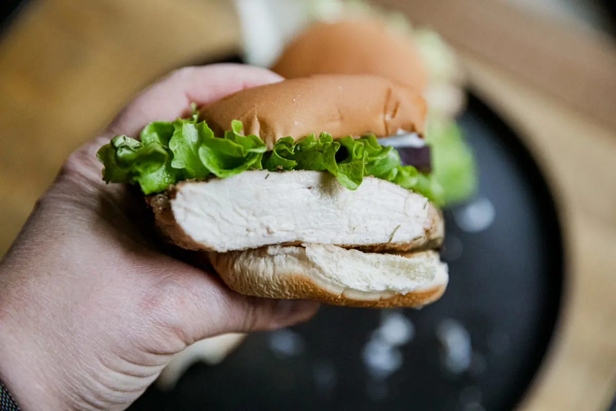 A hand holding a chicken sandwich with lettuce and a slice of tomato visible inside a bun.