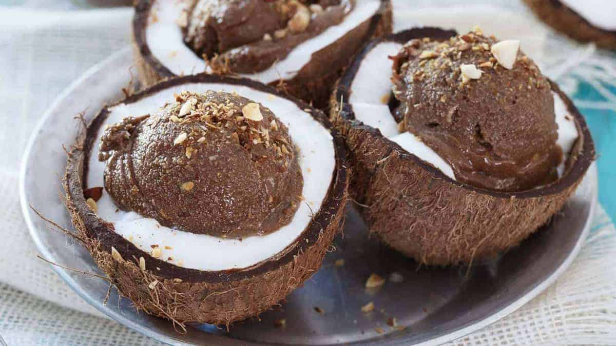 Three halved coconut shells filled with white coconut flesh and topped with scoops of chocolate ice cream and sprinkled chopped nuts on a metal plate.