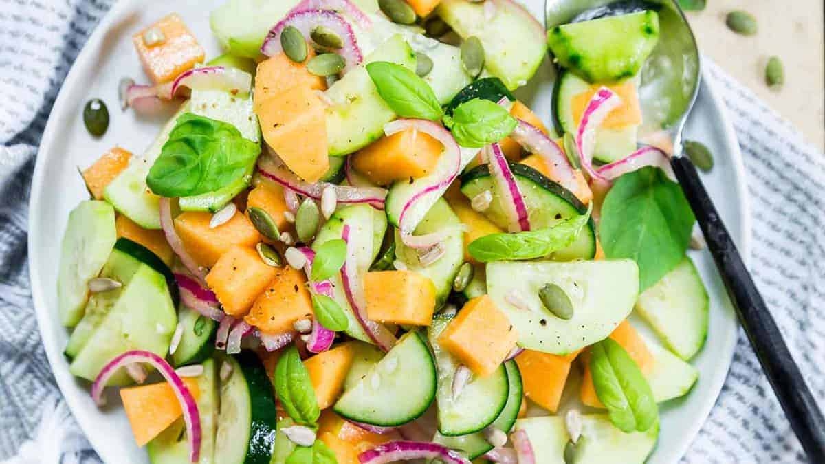 A plate of salad with cucumber slices, cantaloupe cubes, red onion, basil leaves, and seeds, served with a spoon.
