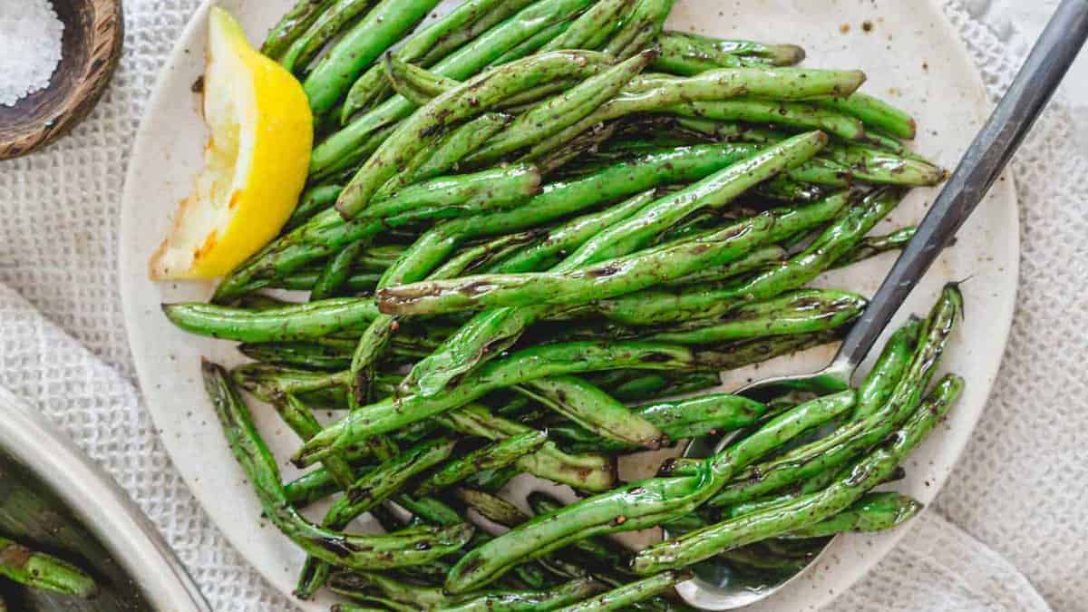 A plate of cooked green beans with grill marks, garnished with a lemon wedge, and a serving spoon on the side.