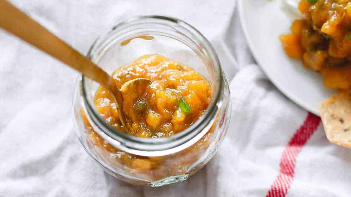 A glass jar filled with orange-colored chutney and a spoon inside, placed on a white cloth next to a plate with more chutney and chips.