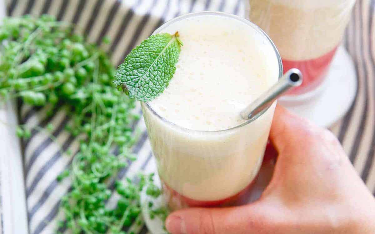 A hand holds a glass of layered drink with a frothy top, garnished with a mint leaf and served with a metal straw. Green herbs and another drink are in the background.