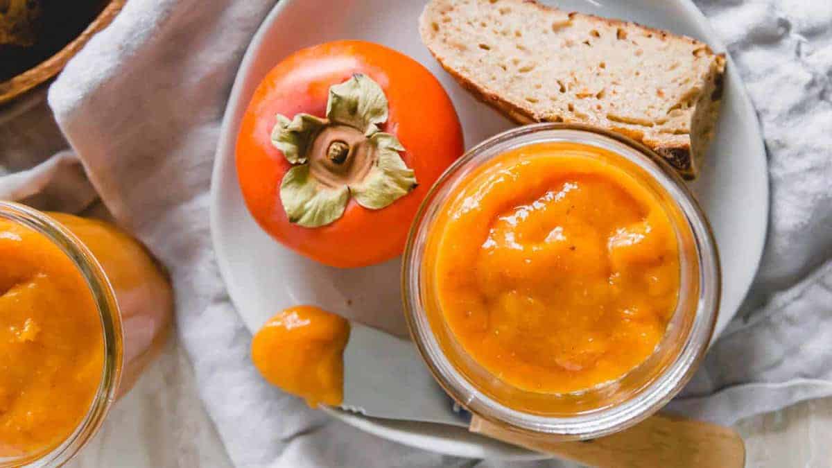 A plate with a whole persimmon, a slice of bread, and a jar of persimmon jam, shown from above on a light-colored cloth.