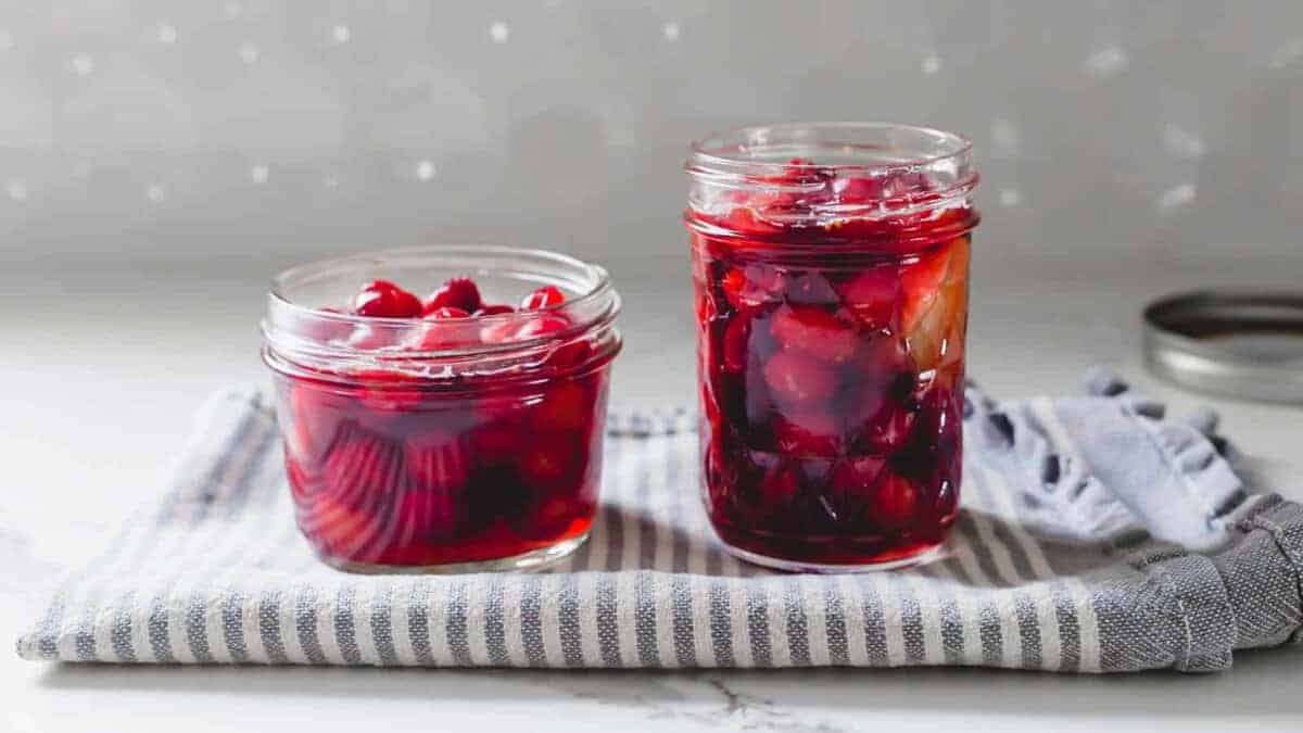 Two glass jars filled with preserved cherries in syrup sit on a striped towel, with a jar lid off to the side.