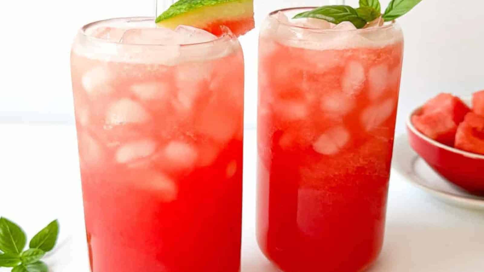 Two tall glasses filled with ice and red watermelon drink, each garnished with a watermelon slice; basil leaves and a bowl of watermelon in the background.
