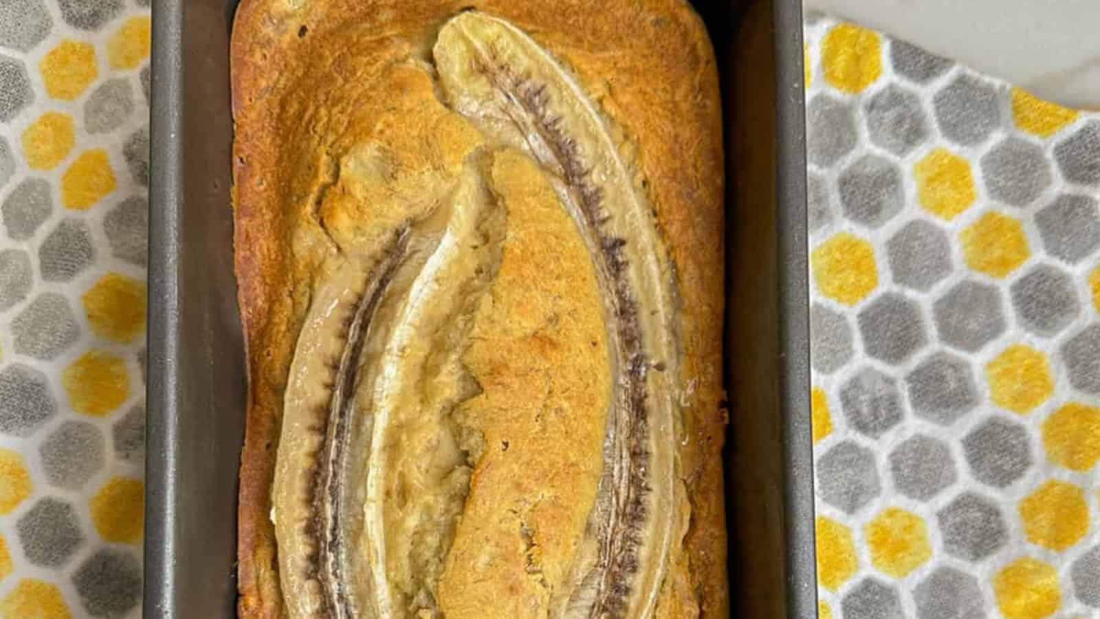Banana bread in a loaf pan, topped with a sliced banana, on a countertop with a hexagonal-patterned towel.