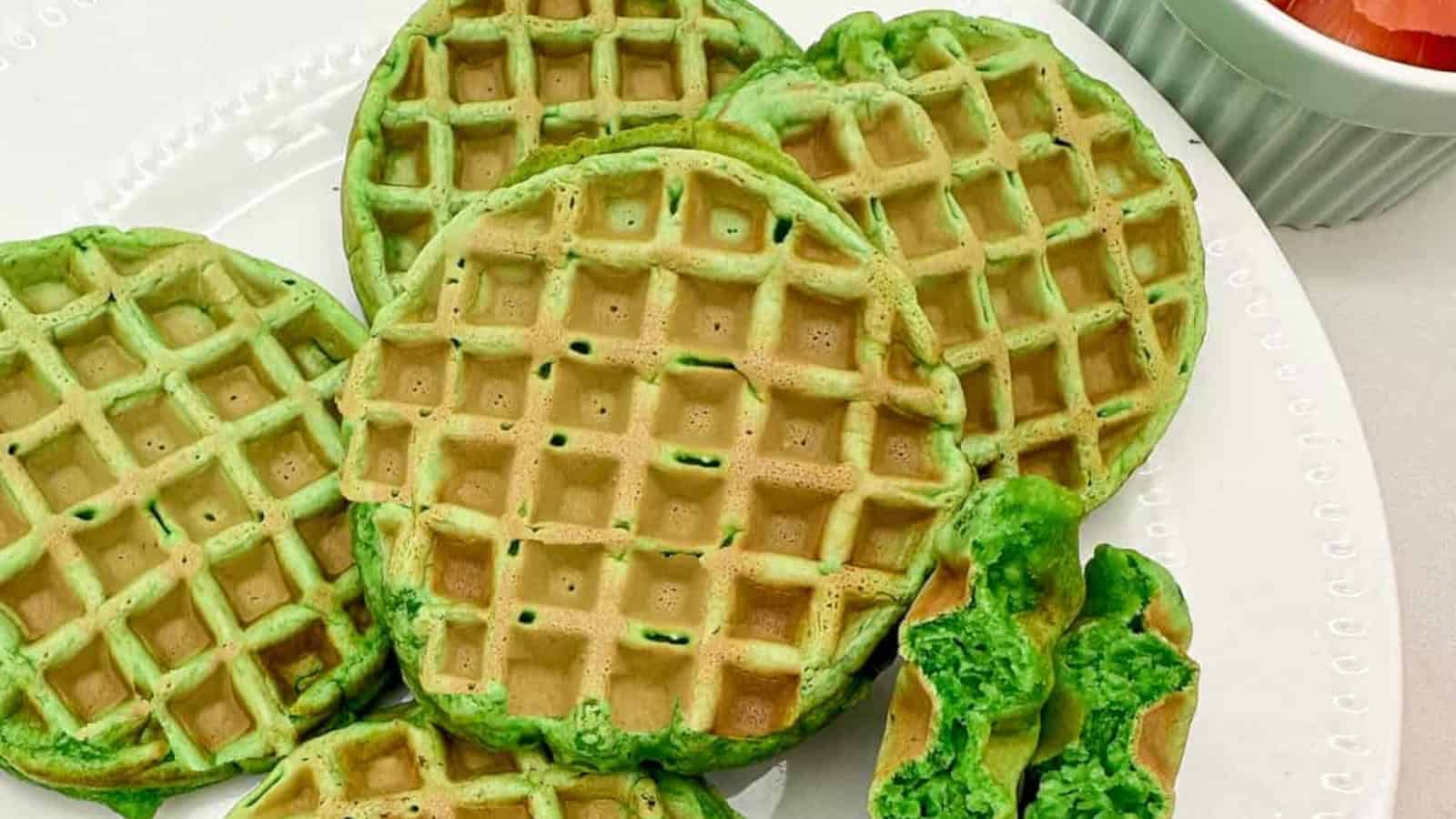 Five green waffles are arranged on a white plate, with one waffle broken in half to show the green interior. A small dish with sliced fruit is partially visible in the corner.