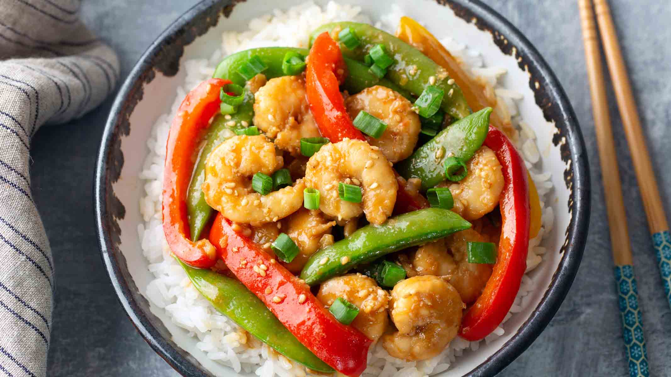 A bowl of rice with shrimp, pea pods, and bell pepper stir fry on top.