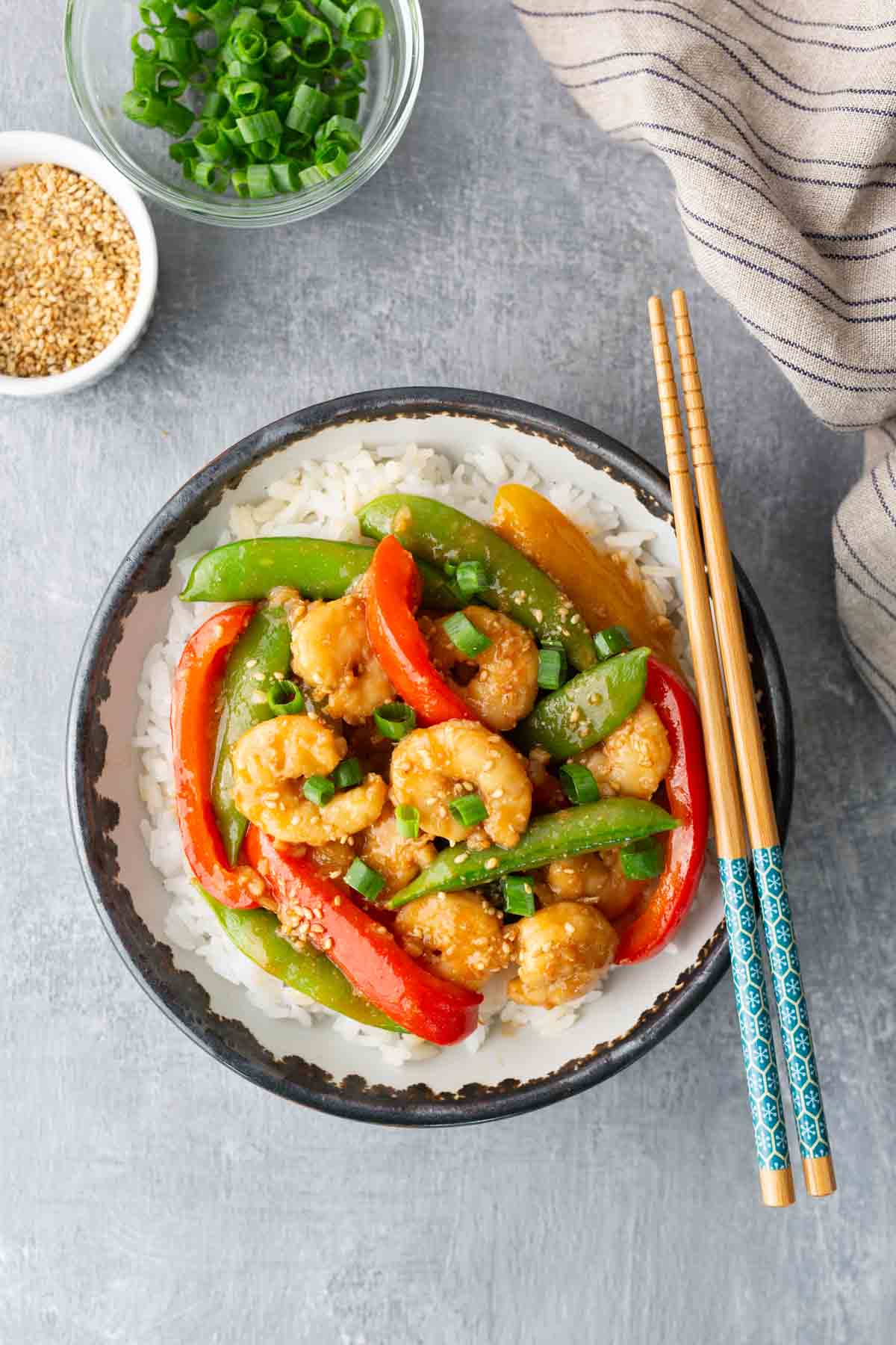 A bowl of white rice topped with shrimp, bell peppers, and snap peas, garnished with green onions, with chopsticks resting on the bowl. Small bowls of sesame seeds and green onions nearby.