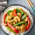 A bowl of white rice topped with shrimp, sliced red bell peppers, snap peas, and chopped green onions, with chopsticks placed nearby.