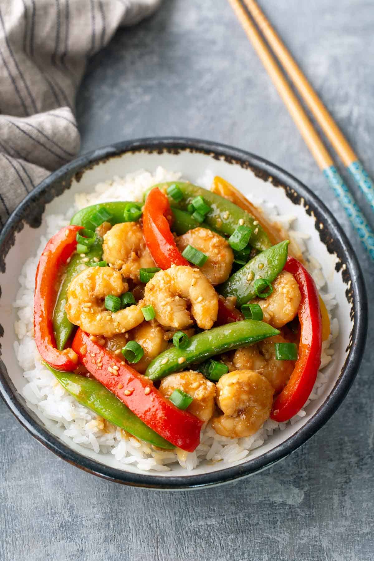 A bowl of white rice topped with shrimp, sliced red bell peppers, snap peas, and chopped green onions, with chopsticks placed nearby.