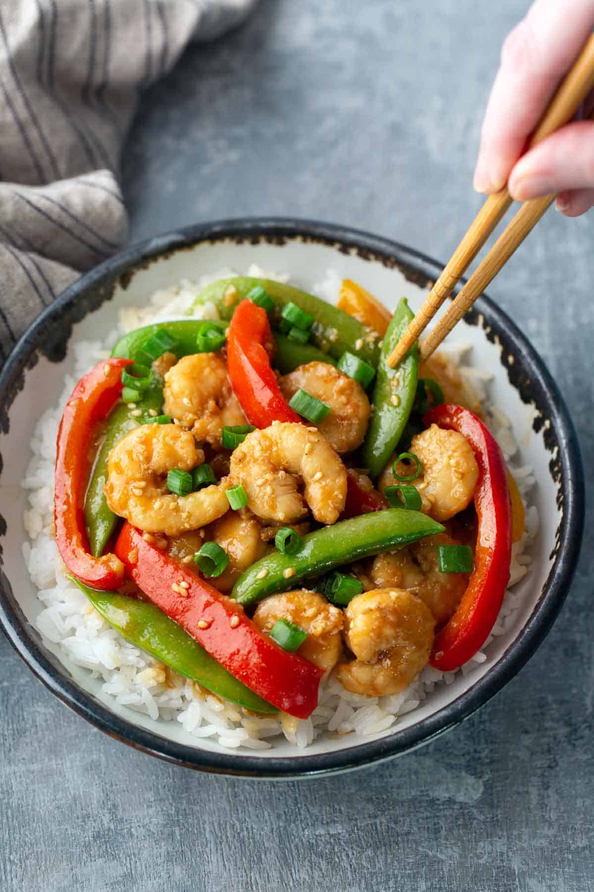 A bowl of white rice topped with shrimp, red bell peppers, snap peas, and chopped green onions; a hand uses chopsticks to pick up a snap pea.