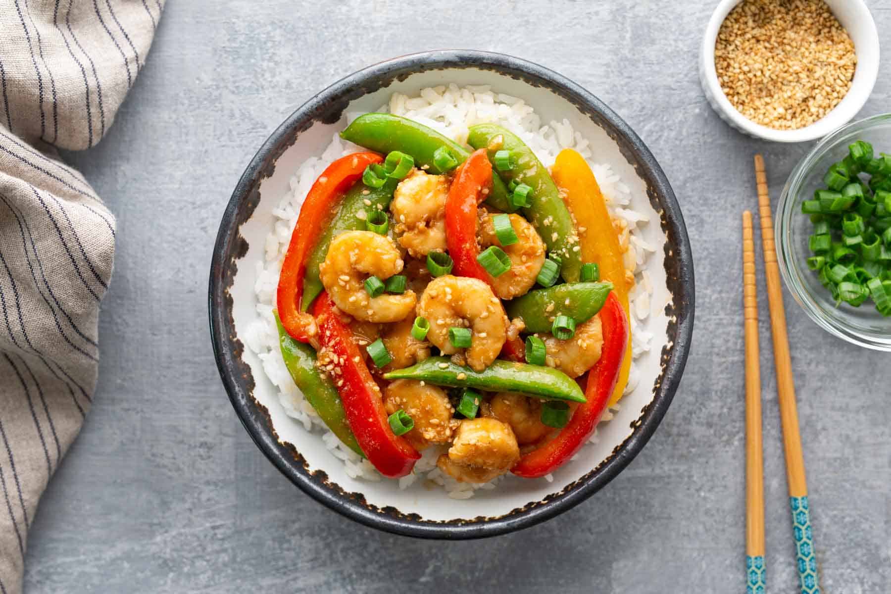 A bowl of rice topped with shrimp stir-fry, including red and yellow bell peppers and snap peas, garnished with chopped green onions. Chopsticks, sesame seeds, and green onions are nearby.