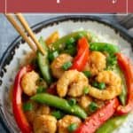 A bowl of shrimp stir fry with red and green bell peppers and snap peas served over white rice, being picked up with chopsticks.
