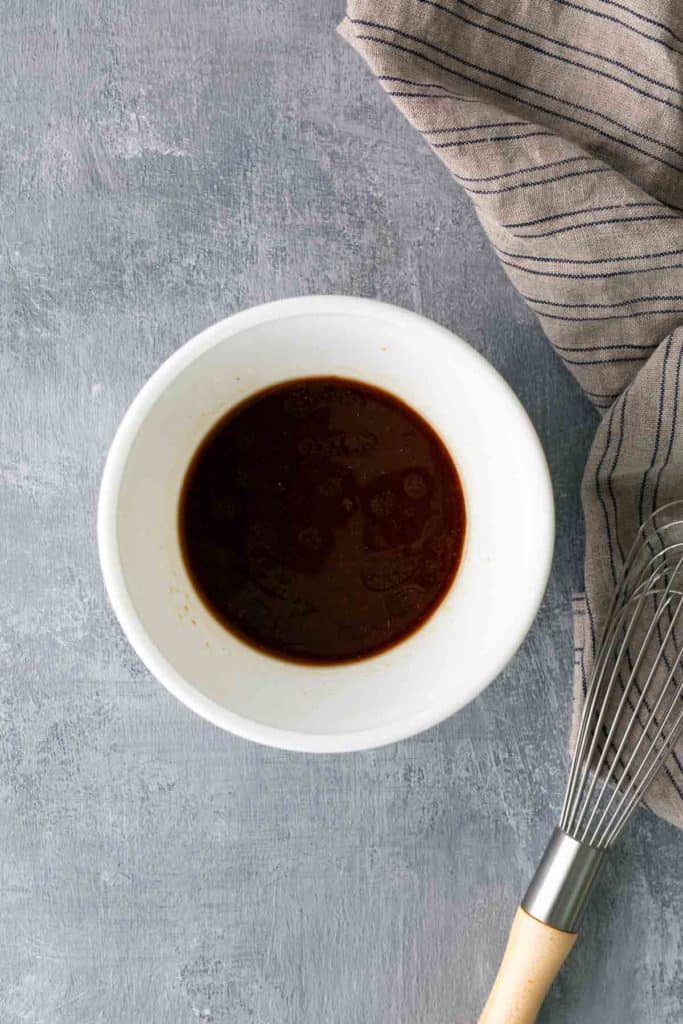 A white bowl containing dark brown liquid sits on a gray surface next to a striped cloth and a metal whisk with a wooden handle.