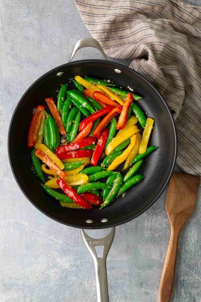 Sautéed red, yellow, and green bell peppers with snap peas in a black skillet, placed on a gray surface next to a wooden spatula and striped cloth.